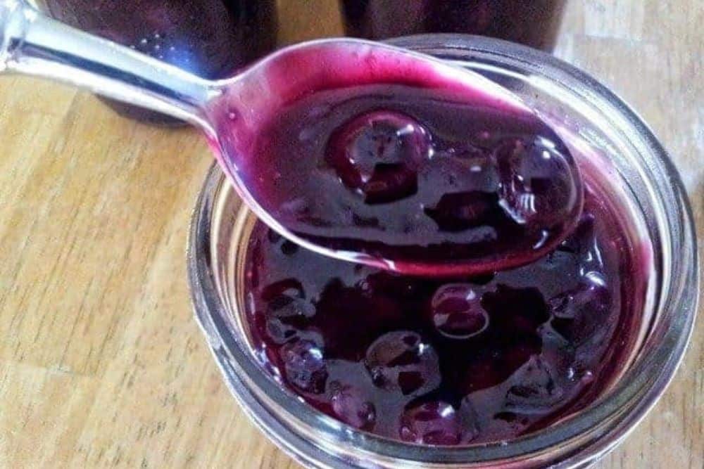 A spoon filled with dark purple jam held above a glass jar filled with the same jam. The jar is placed on a wooden surface.