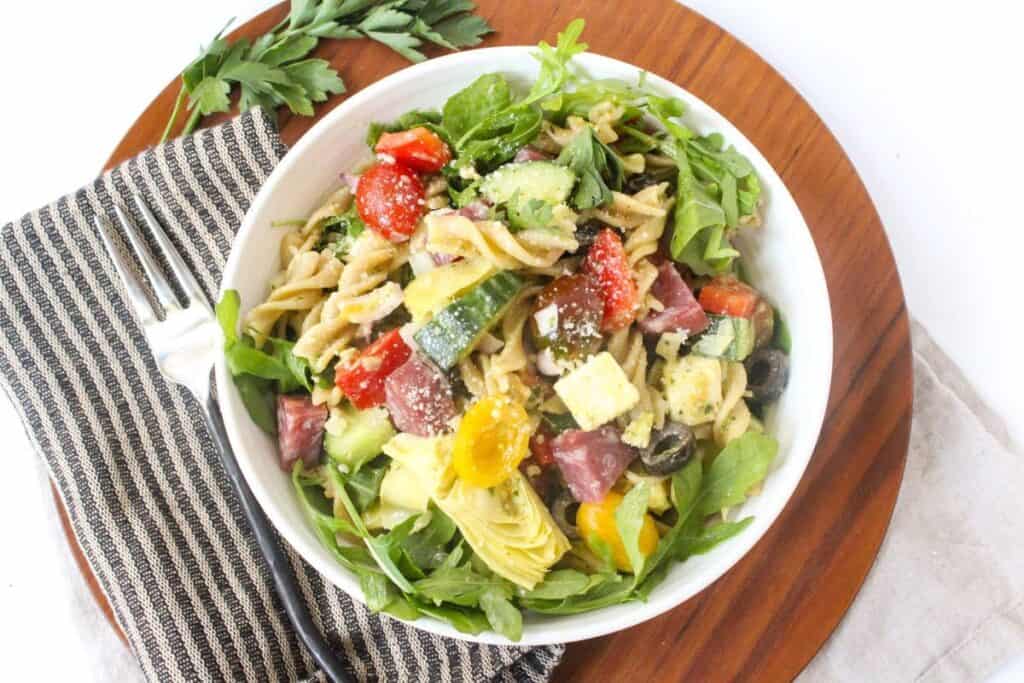 A bowl of pasta salad with mixed vegetables including tomatoes, cucumbers, olives, and arugula, served on a striped cloth next to a fork and parsley garnish.