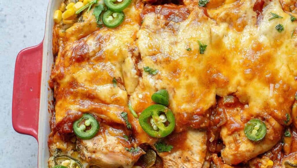 A close-up of a cheesy, baked casserole topped with sliced jalapeños, corn, and melted cheese. The dish is in a red baking tray.
