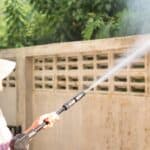 A person in a hat uses a pressure washer to clean a concrete wall outdoors.