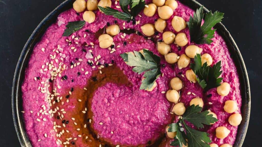 A bowl of bright pink beet hummus garnished with whole chickpeas, parsley leaves, sesame seeds, and a swirl of oil on the surface.