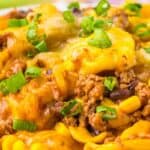 A close-up of a cheesy taco pasta dish topped with ground beef, beans, corn, melted cheese, and chopped green onions.