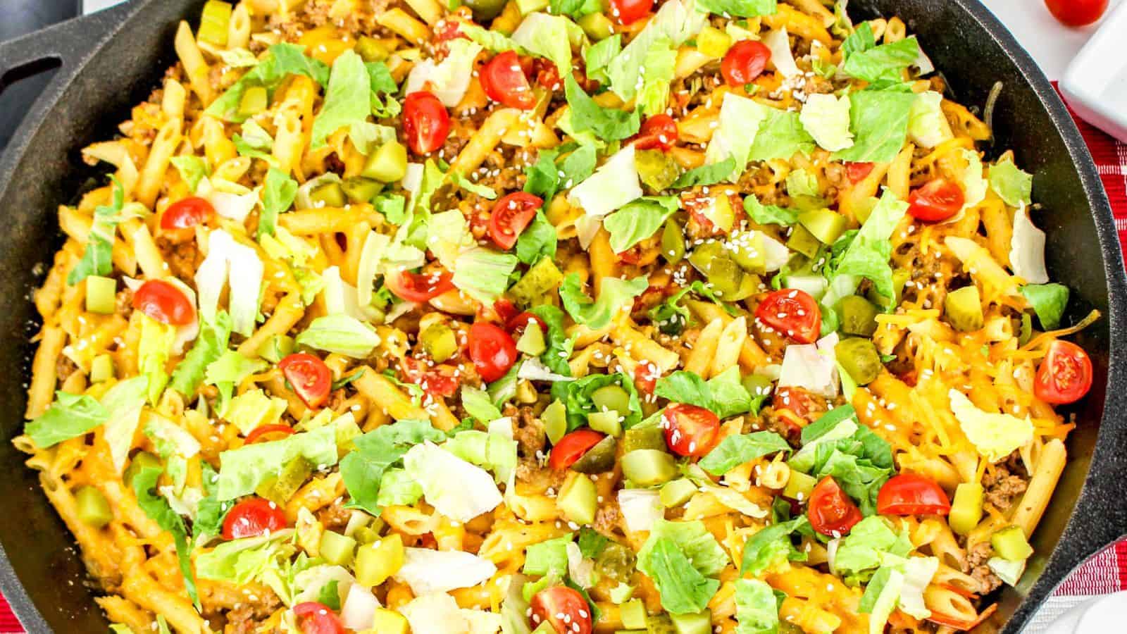 A skillet filled with penne pasta topped with shredded cheese, ground beef, diced tomatoes, lettuce, and pickles—a delicious take on cheeseburger pasta.