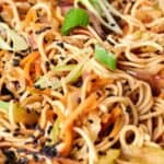 A close-up of a plate of stir-fried noodles with vegetables, garnished with black sesame seeds and green onions. Chopsticks are visible on the left side of the plate.