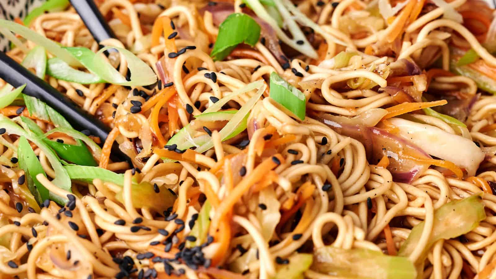 A close-up view of a plate of chow mein stir-fried noodles with vegetables and black sesame seeds, with chopsticks visibly picking up a portion.