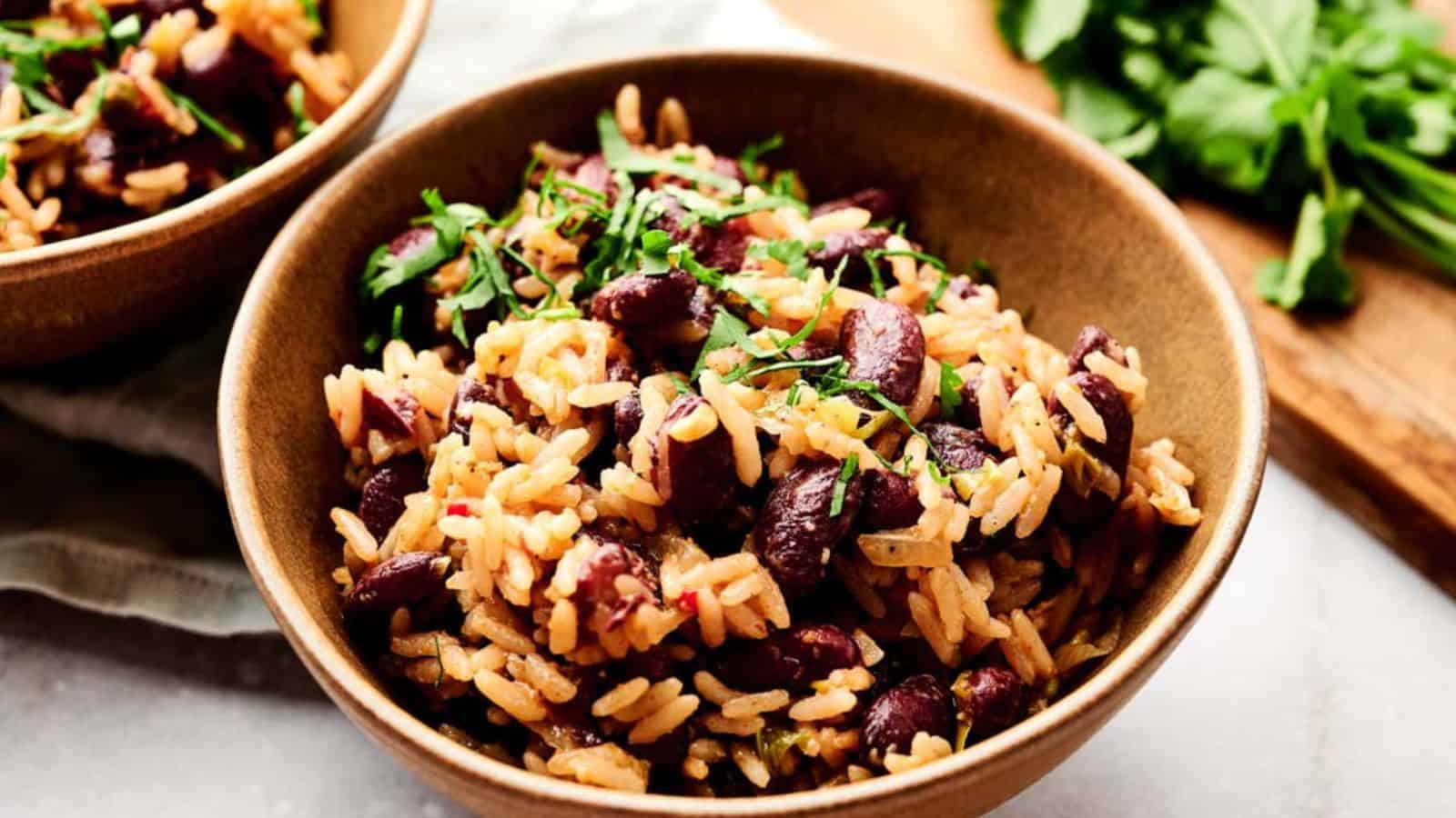 A bowl of cooked rice mixed with kidney beans, garnished with chopped herbs on top, placed on a light-colored surface with fresh greens on a wooden board in the background.