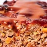 Close-up of a sliced meatloaf topped with a glossy glaze. Visible ingredients include ground meat, diced vegetables, and seasonings.