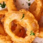 Close-up of golden, crispy onion rings garnished with parsley and black pepper on a white surface.