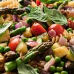 A plate featuring a colorful pasta salad with fusilli, cherry tomatoes, asparagus, peas, red onion, zucchini, and fresh basil leaves.