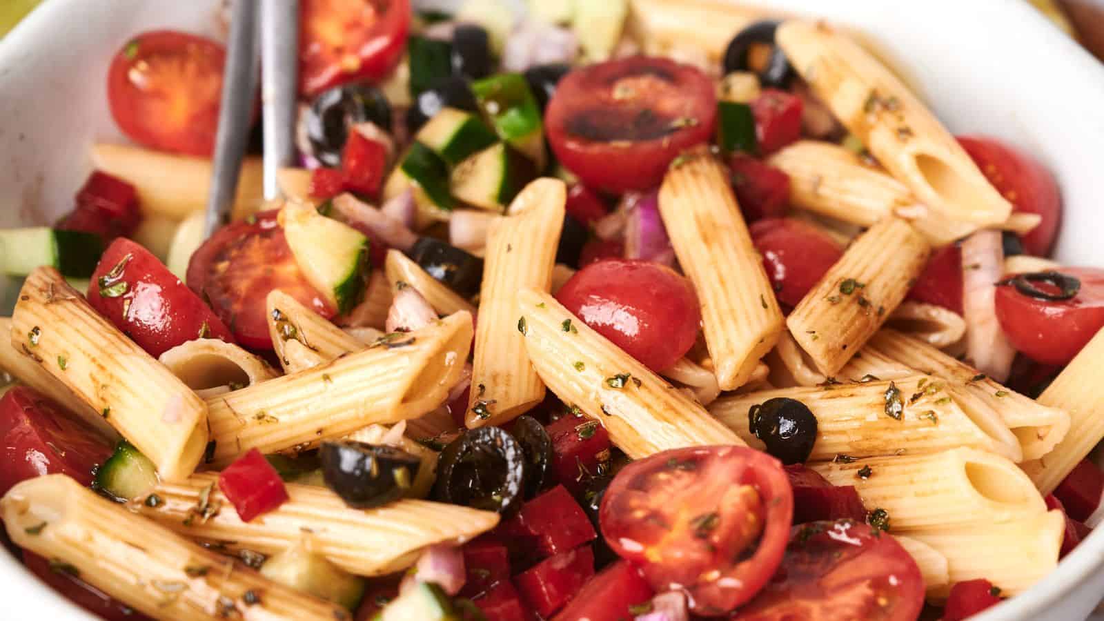 A bowl of vibrant pasta salad with penne, cherry tomatoes, black olives, diced cucumbers, red onions, and a drizzle of dressing.
