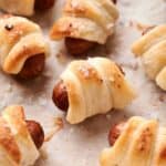 Close-up of multiple pigs in a blanket, which are small sausages wrapped in golden, flaky pastry on a parchment-lined surface, sprinkled with coarse salt.