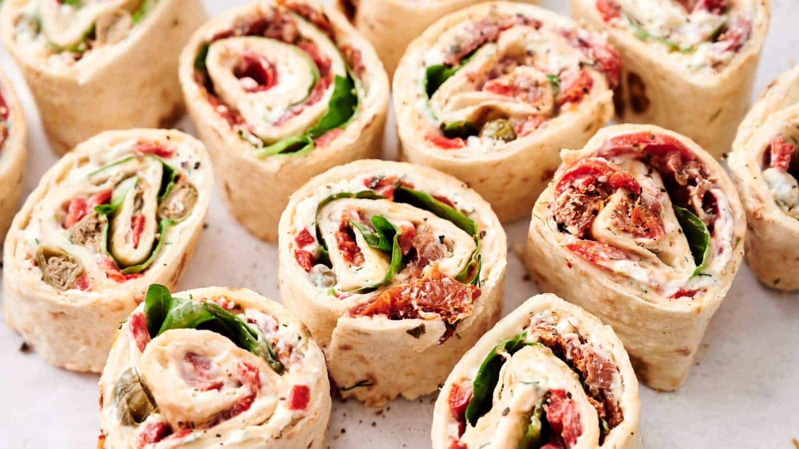 Close-up of several tortilla pinwheel wraps filled with various ingredients including spinach, cream cheese, tomatoes, and other vegetables, arranged neatly on a white surface.