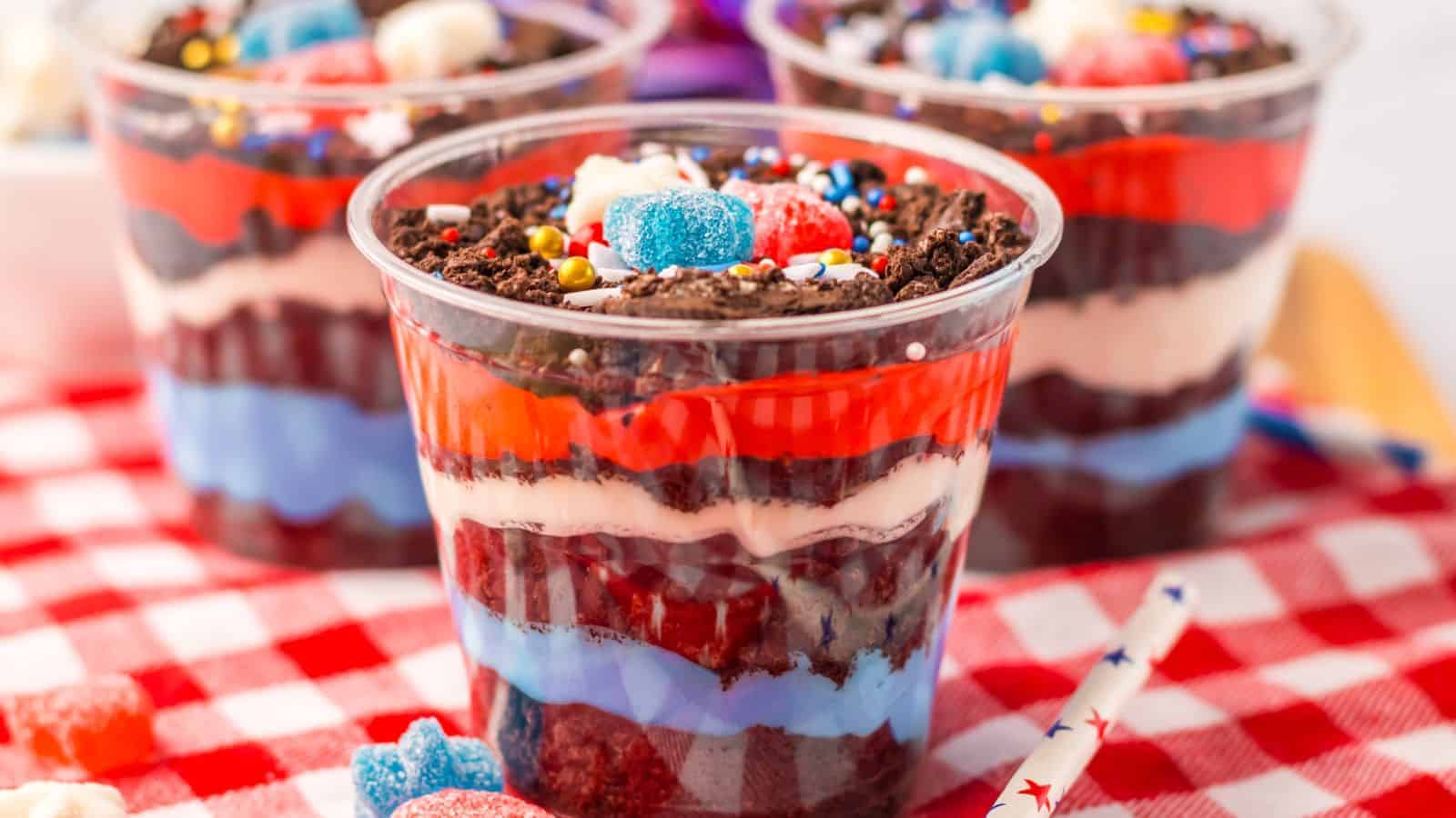 Three-layered red, white, and blue dirtcups with vibrant fillings are topped with various candies and sprinkles, all perfectly arranged on a red and white checkered cloth.