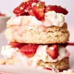 A strawberry shortcake on a plate, featuring two layers of biscuit, fresh whipped cream, and sliced strawberries on top, with a bowl of more strawberries in the background.