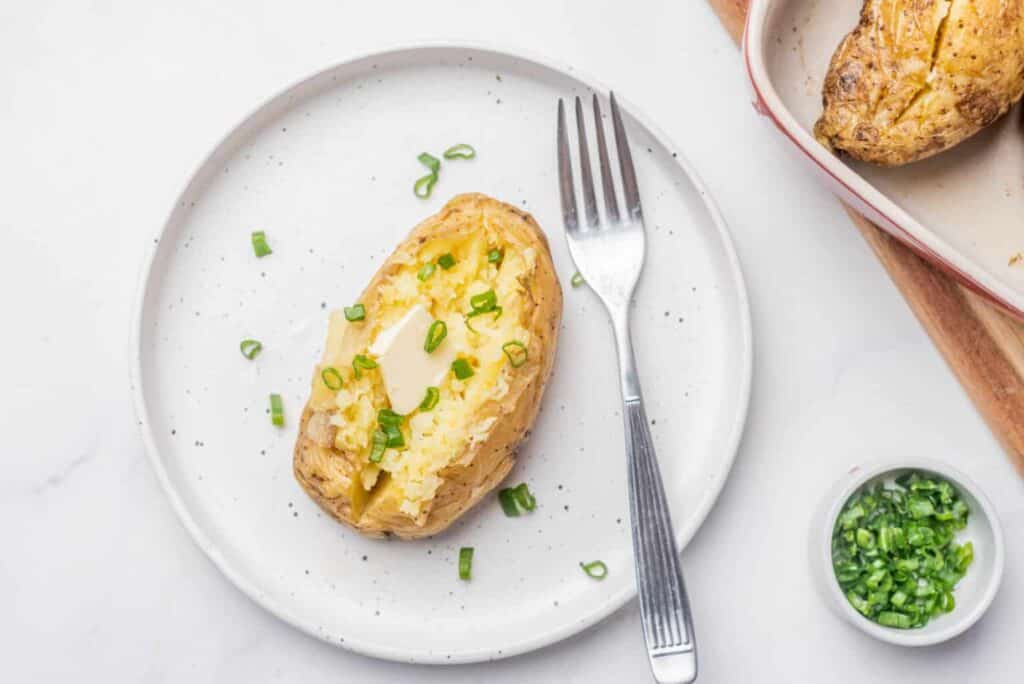 A baked potato topped with melted butter and chopped green onions sits on a white plate with a fork. Nearby, a dish with another baked potato and a small bowl of chopped green onions are visible.