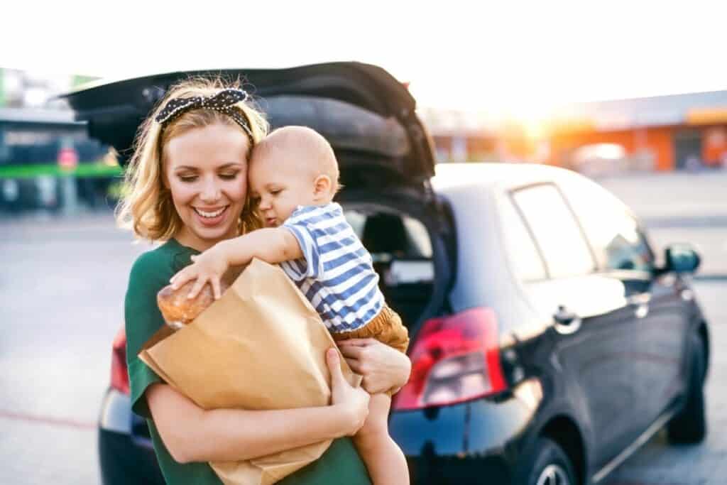 A woman holds a child and a paper bag of groceries near an open car trunk in a parking lot.