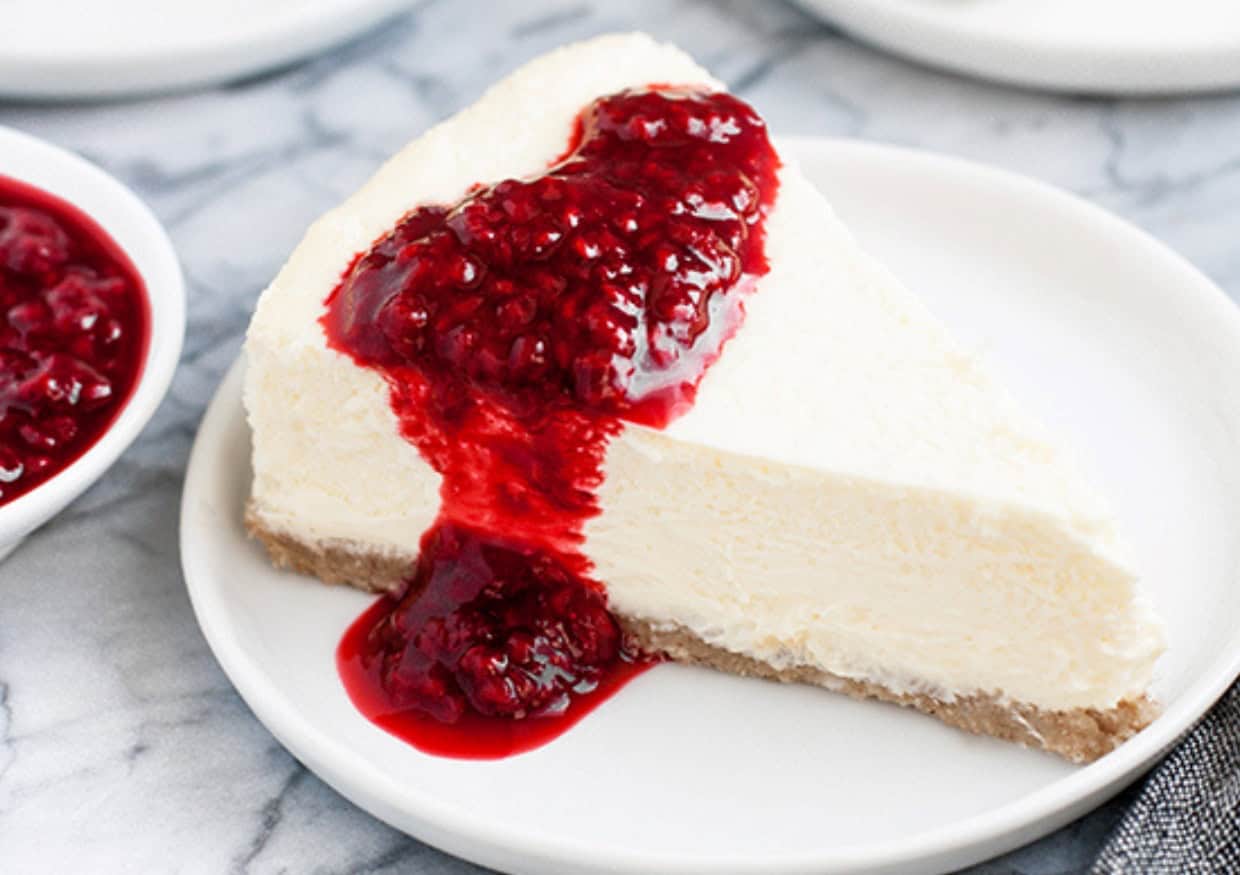 A slice of cheesecake topped with raspberry sauce, served on a white plate, with a small bowl of additional raspberry sauce nearby.