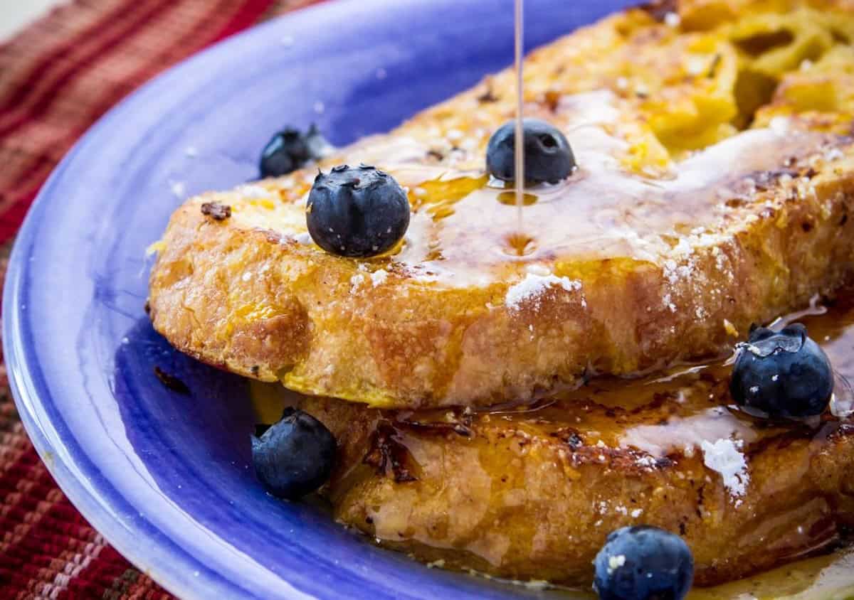 A plate with two slices of French toast topped with blueberries and being drizzled with syrup.