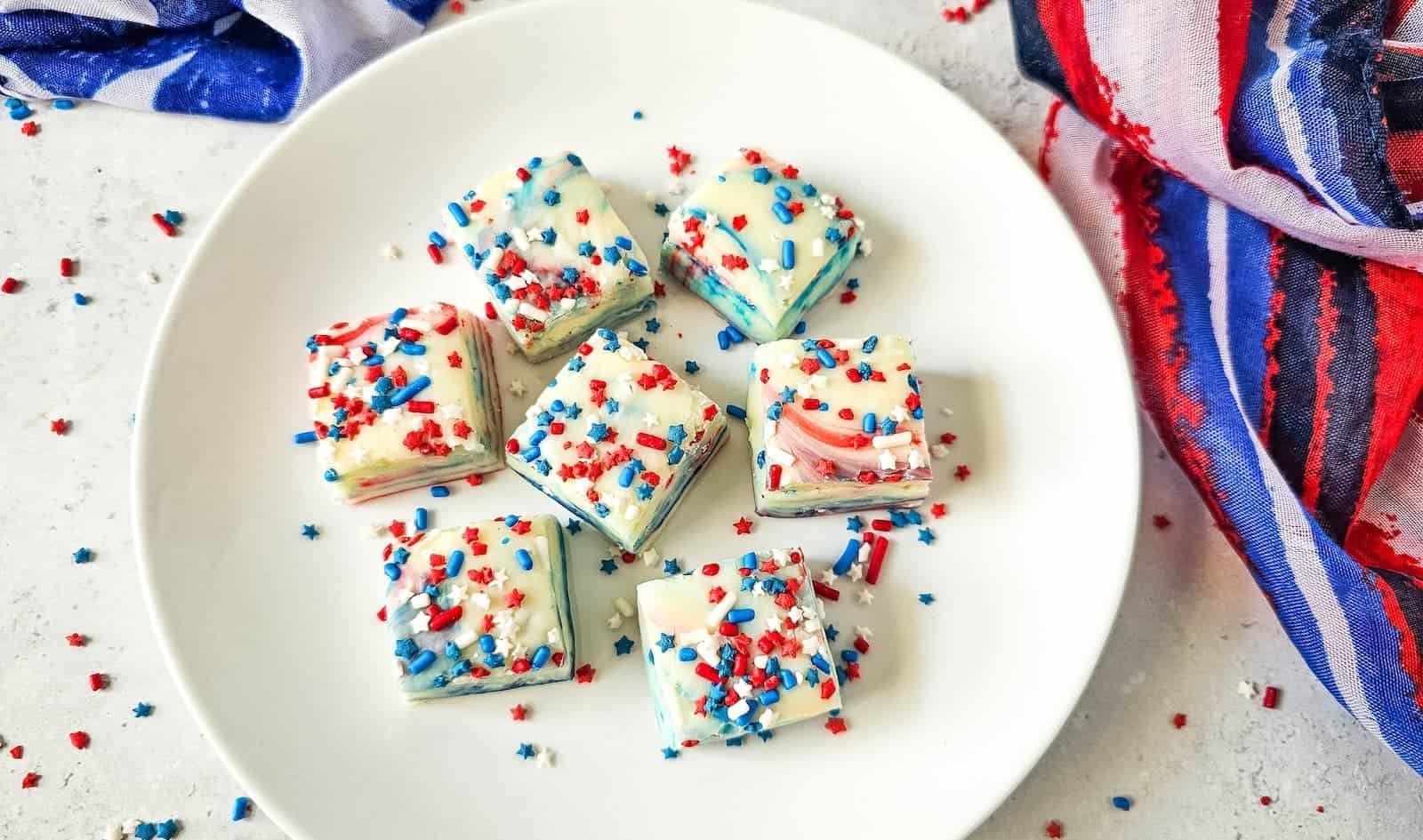 A white plate holds seven pieces of fudge decorated with red, white, and blue sprinkles, situated on a white surface with a red, white, and blue cloth nearby.