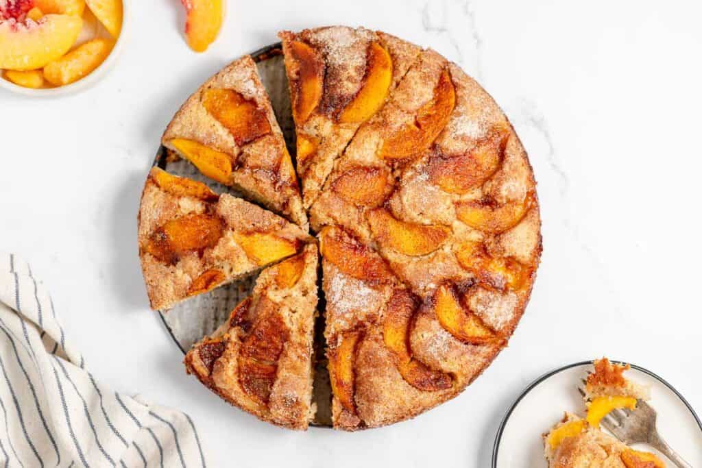 A round, sliced peach cobbler cake on a plate, next to a small bowl of peach slices and a striped cloth. A slice of the cake is on a separate plate with a fork.