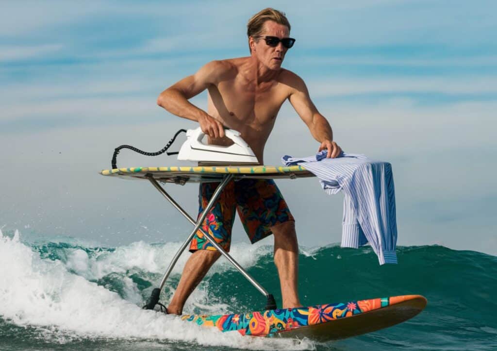 A man with sunglasses surfs on a wave while ironing a shirt on an ironing board.