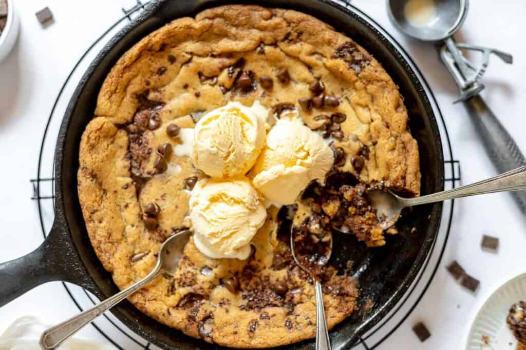 A skillet cookie topped with three scoops of vanilla ice cream is being shared with three spoons. Cookie pieces and chocolate chips are visible, alongside a small dish and ice cream scoop.