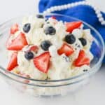 A glass bowl filled with a dessert mixture of whipped cream, sliced strawberries, blueberries, and small marshmallows, placed on a white surface with a blue cloth and additional strawberries nearby.