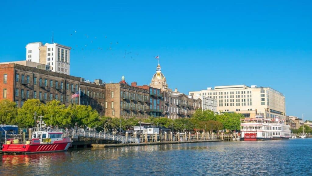 Waterfront scene showing historic buildings along the river with boats docked; clear blue sky and flags visible—one of the charming sights you'll encounter while exploring things to do in Savannah with kids.