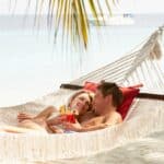 A couple relaxes in a hammock by the beach, holding drinks with straws and garnishes, under a palm tree with a boat visible in the background on the water.