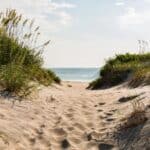 A sandy path between tall grasses leads to a serene beach with calm waters under a partly cloudy sky.