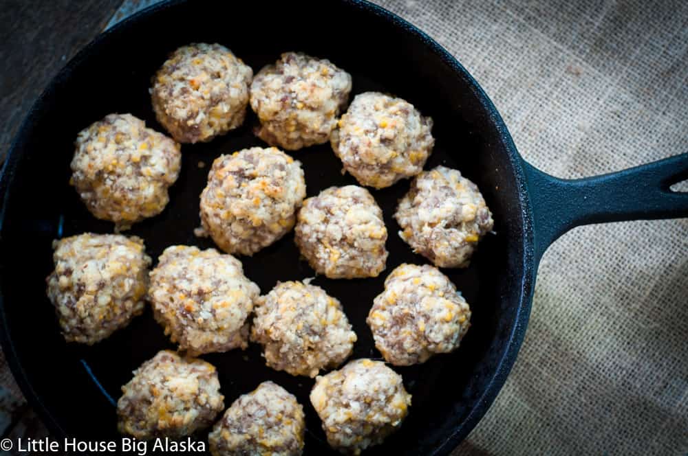 A cast iron skillet containing 15 evenly spaced uncooked sausage balls on a burlap surface.