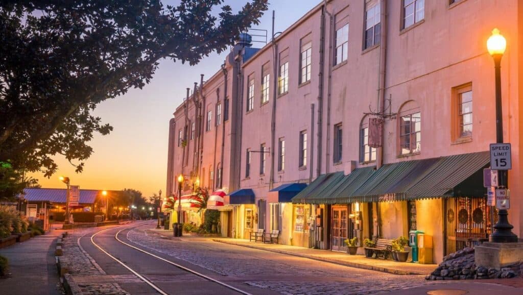 A historic building with shops and awnings along a cobblestone street with tram tracks is illuminated by streetlights at sunset. A speed limit sign reads 15 mph, adding to the timeless charm—a perfect spot for exploring things to do in Savannah with kids.