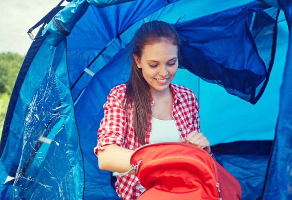 A woman in a red checkered shirt is sitting inside a blue tent, packing or unpacking a red backpack. She is smiling and looks focused on her task.