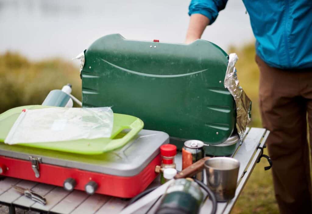 Person setting up a green camping stove on a picnic table with various kitchen items, including a red stove, green cutting board, canned food, and a mug. Outdoor scenery in background.