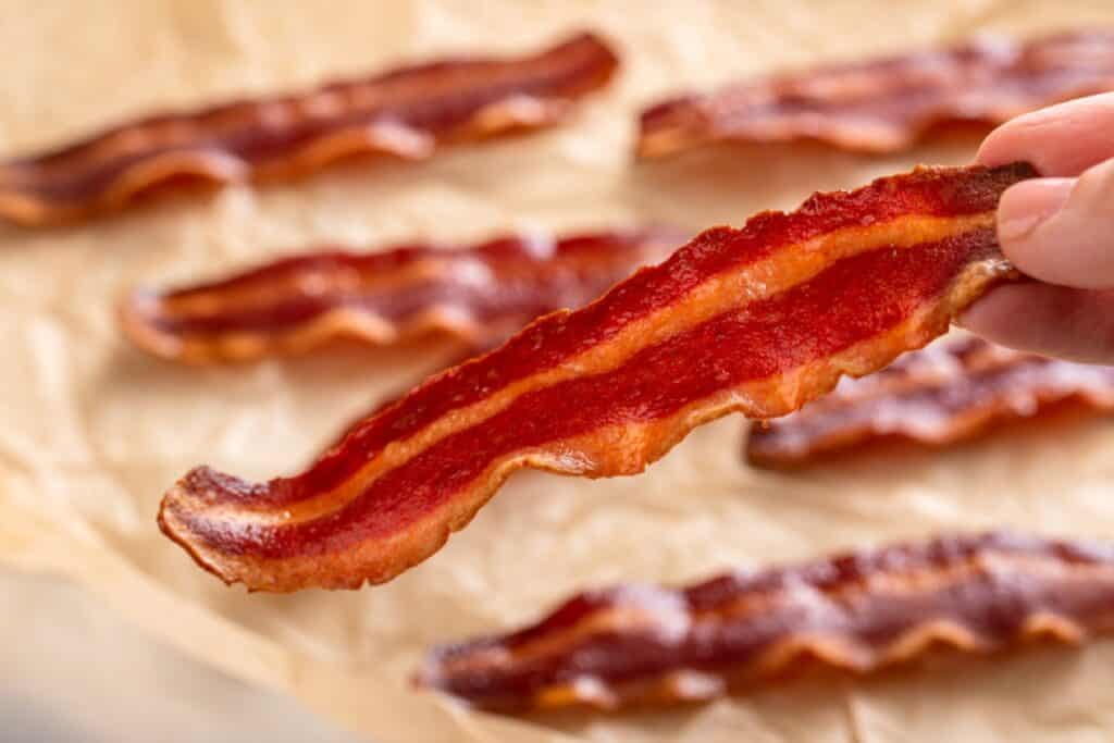 Close-up of a hand holding a crispy strip of bacon with more bacon strips on parchment paper in the background.