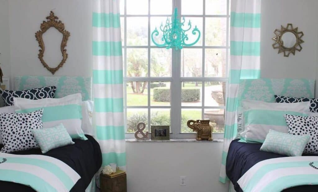 A bedroom with two twin beds featuring teal and white bedding, striped teal and white curtains, a large window in the center, and various decor items including pillows, frames, and a chandelier.