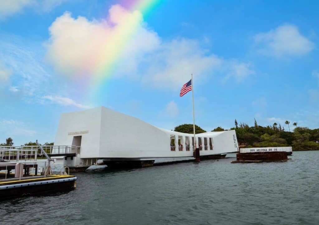 A white memorial stretches across the water in Pearl Harbor as a rainbow shines down from the blue sky above.