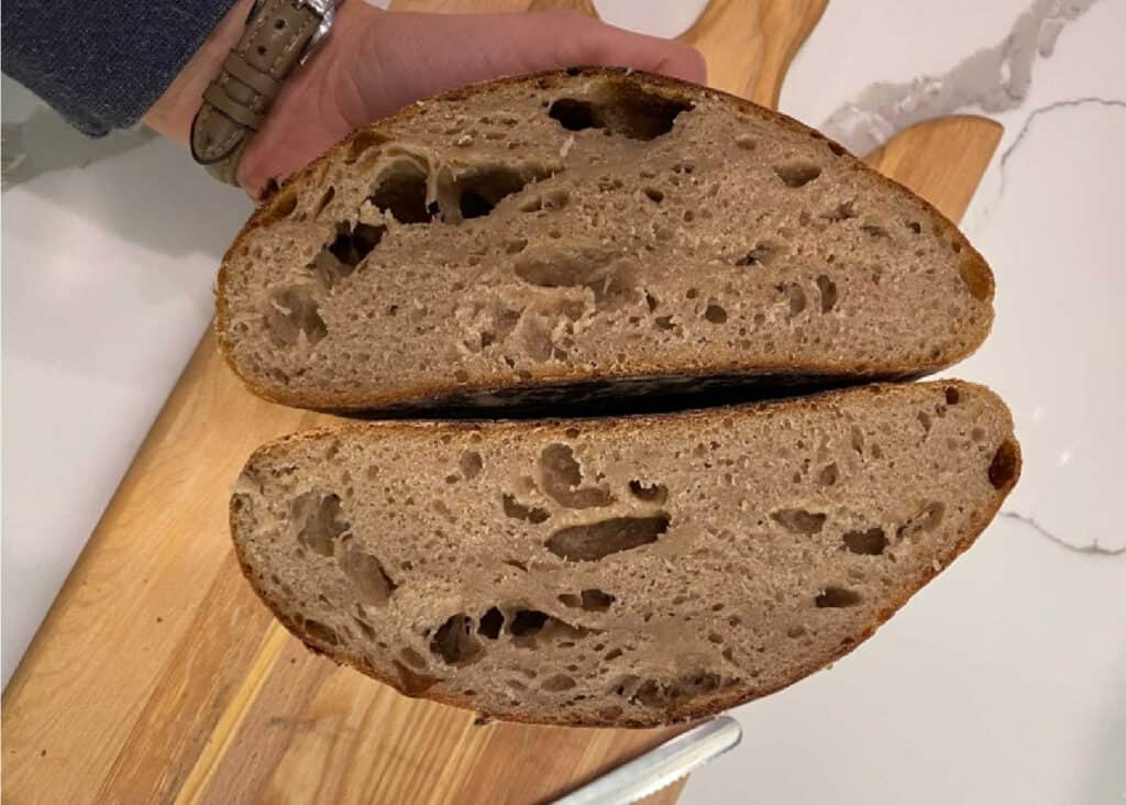A person holding two halves of a loaf of bread with a porous interior on a wooden cutting board.