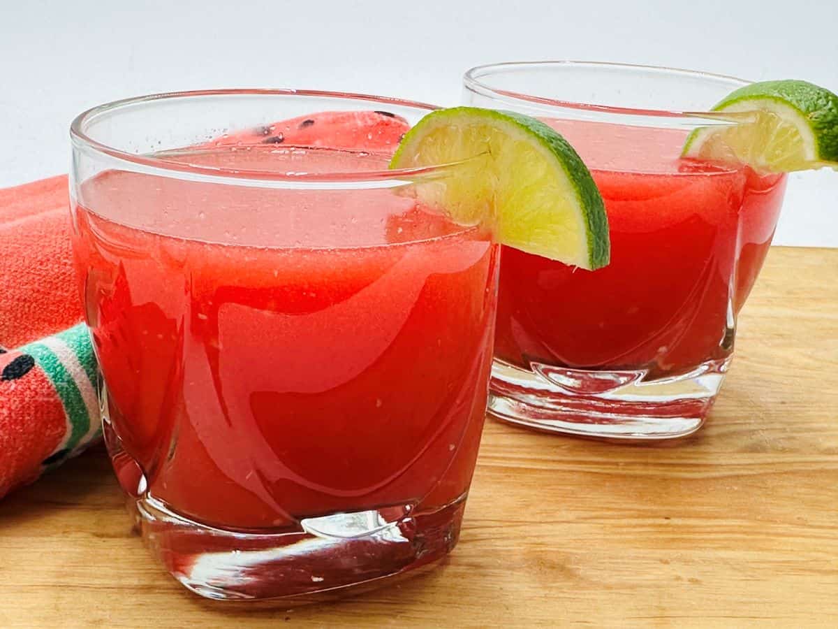 Two glasses of watermelon juice garnished with lime slices are on a wooden surface with a red napkin in the background.