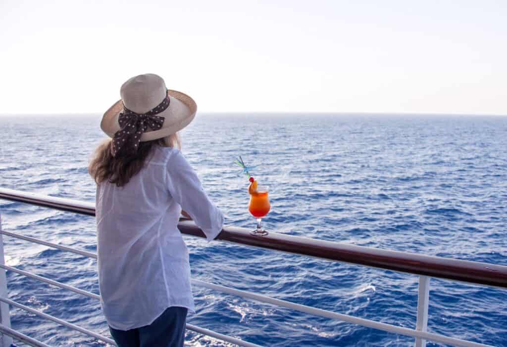 A person wearing a sunhat and white shirt looks out over the ocean from a ship's railing, with a tropical drink placed on the rail next to them. For those seeking cruise tips, this serene moment encapsulates the relaxation awaiting you on your next adventure.