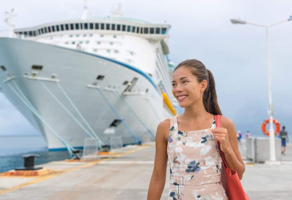 A woman in a floral dress smiles while standing on a pier, with a large white cruise ship docked in the background, ready to share some helpful cruise tips.