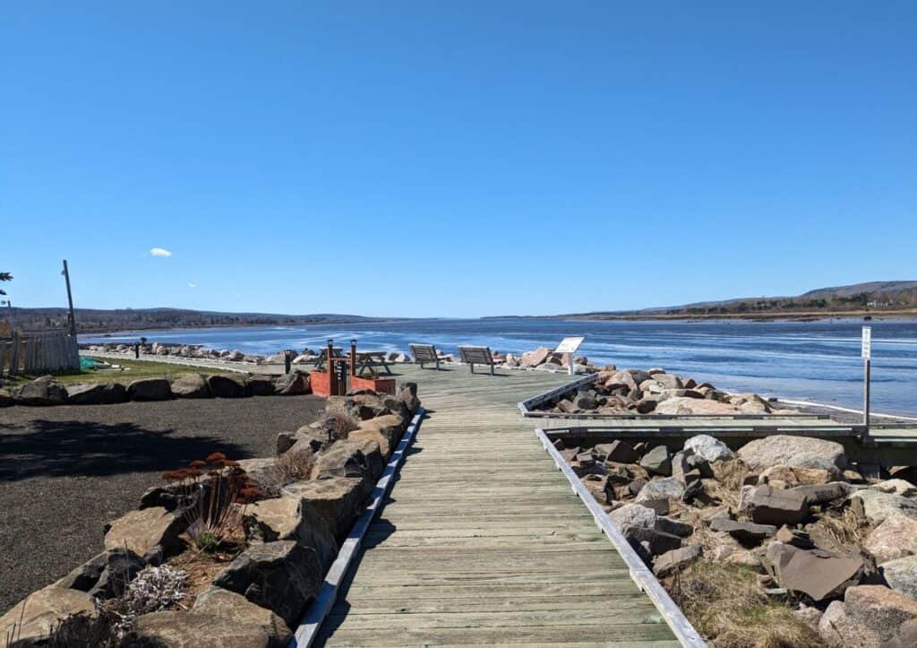 A wooden boardwalk lined with rocks extends to a lake, with chairs at the end. The scene includes clear blue skies, calm water, and distant hills.