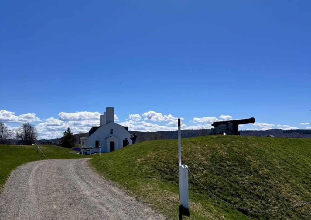 A curved gravel path leads to a white building with a small tower under a clear blue sky. To the right, an old cannon sits on a grassy mound.