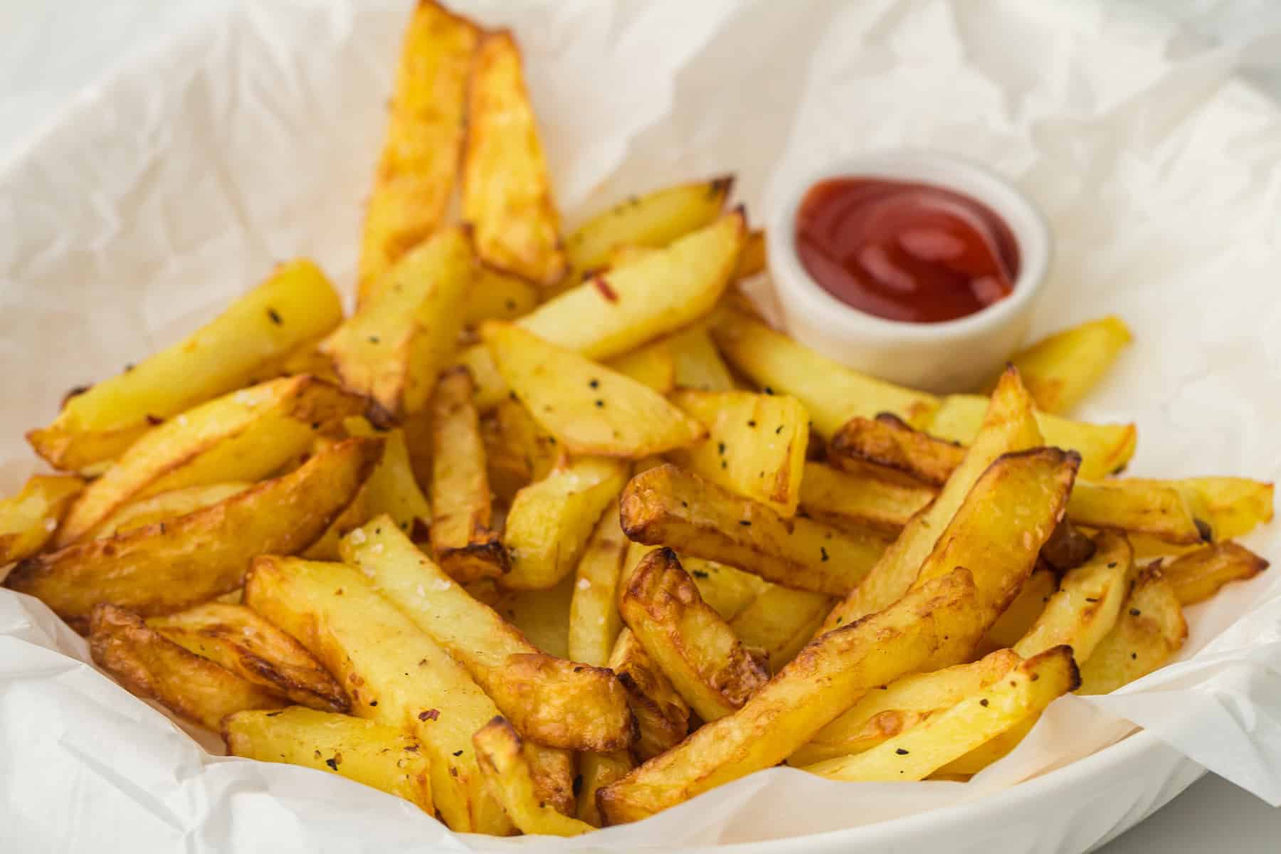 A basket of golden, crispy potato fries seasoned with pepper, accompanied by a small cup of ketchup on the side, presented on white parchment paper.