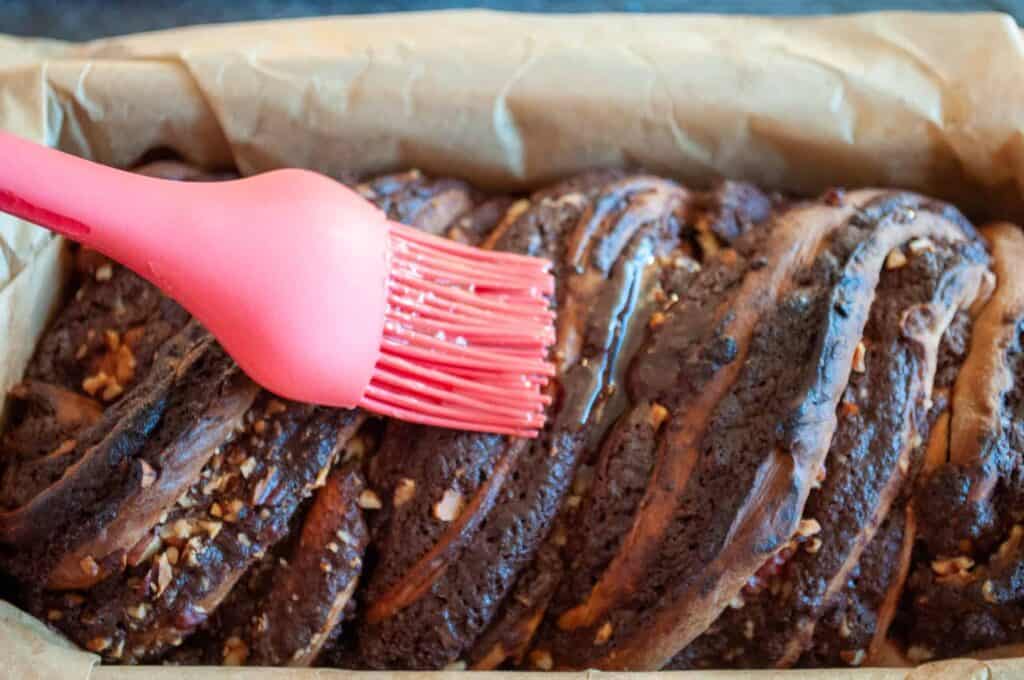 A close-up of a chocolate babka being brushed with glaze using a pink silicone brush. The babka is in a parchment-lined baking pan.