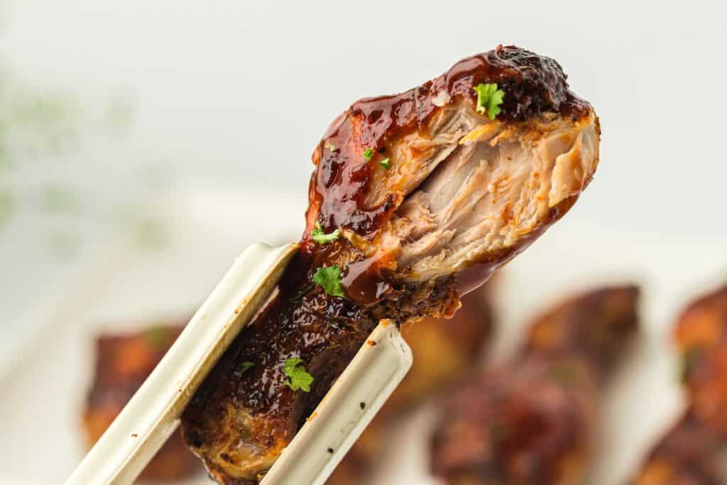 Close-up of a chopstick holding a cooked chicken wing glazed with sauce and garnished with herbs, with more chicken wings in the blurred background.