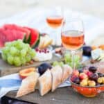 A beach charcuterie board with glasses of rosé wine, a variety of fruits and sliced baguette.
