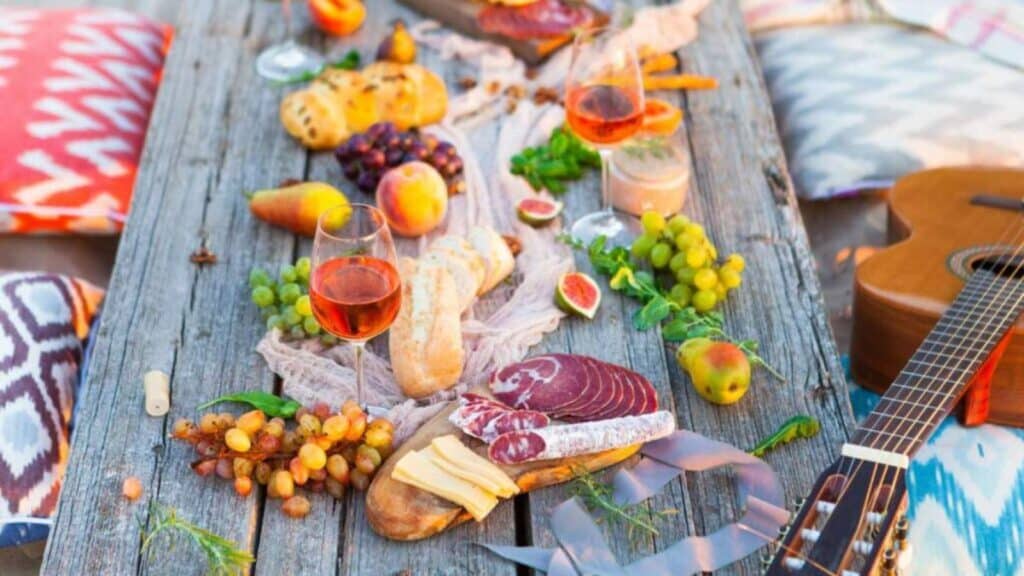 A wooden beach table with assorted food items, including sliced meats, cheese, bread, grapes, figs, and peaches.
