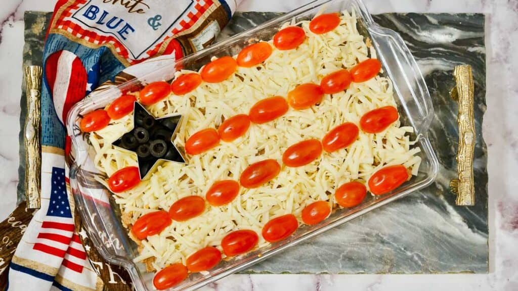 A rectangular dish with a cheese-covered casserole is decorated with cherry tomatoes in horizontal rows and a corner of sliced black olives, resembling an American flag. A patriotic kitchen towel is beside it.