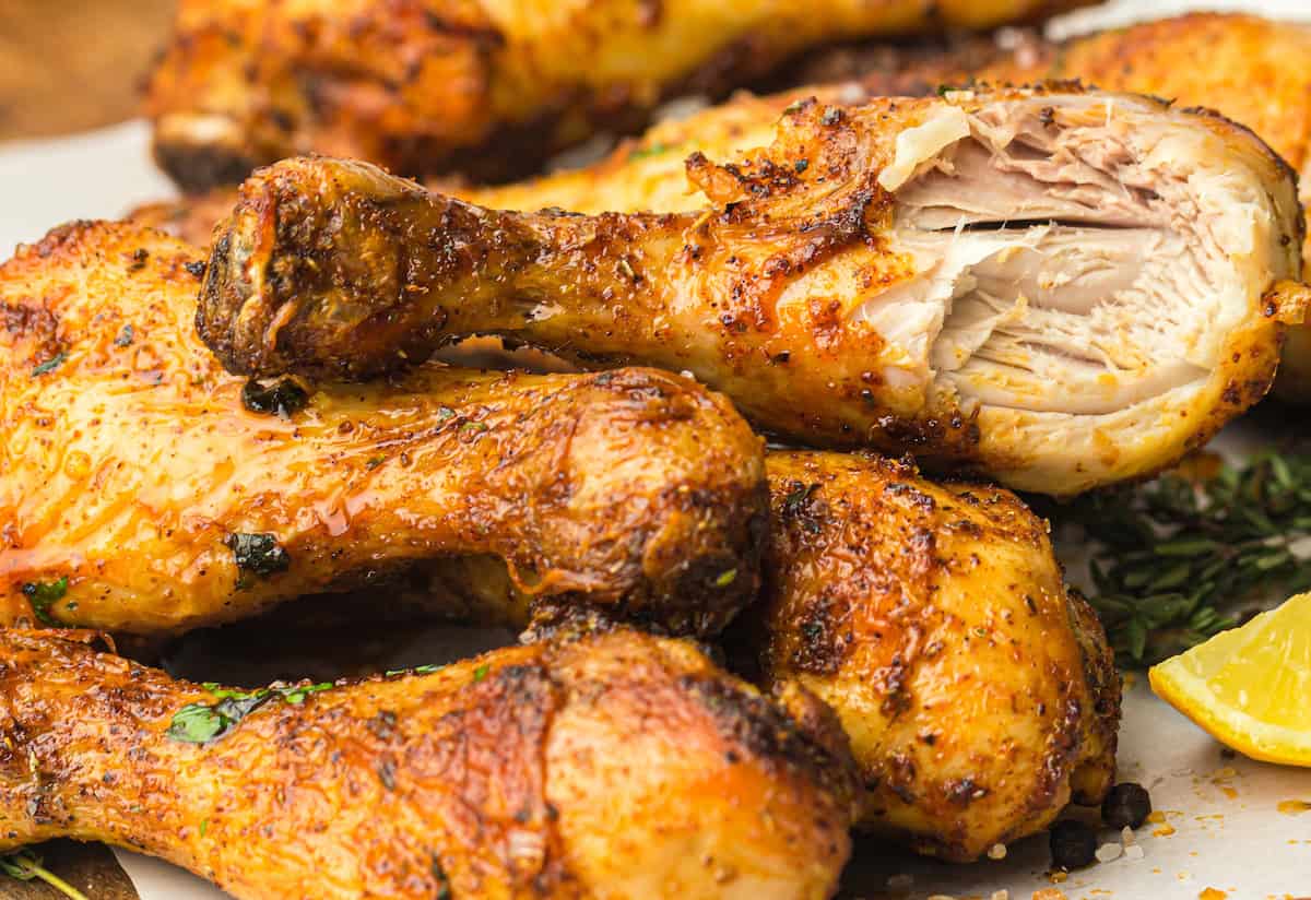 Close-up of a stack of seasoned, grilled chicken drumsticks, with one drumstick partially eaten, revealing moist, cooked meat inside. A lemon wedge and herbs are visible in the background.